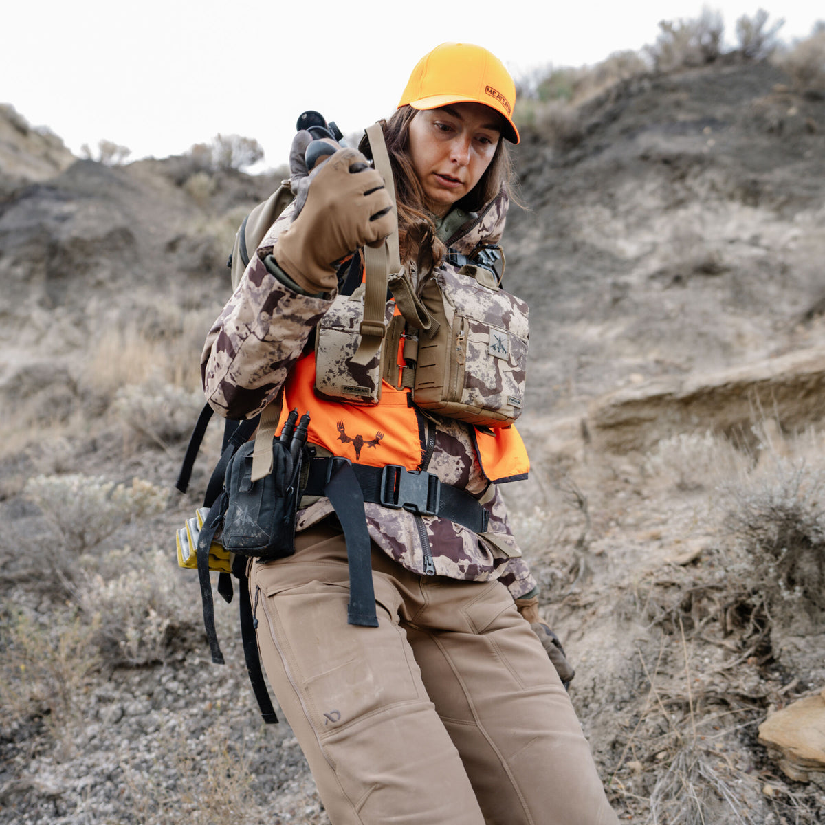 A female hunter wears a brown camouflage jacket, orange vest with a black deer logo, tan pants, tan gloves, and an orange "MEATEATER" cap, holding binoculars.