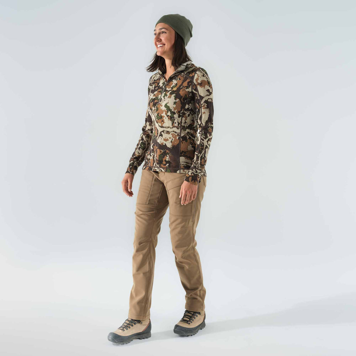 Woman in an olive green beanie, a camouflage 1/4 zip long-sleeve top, tan hiking pants, and tan hiking boots.