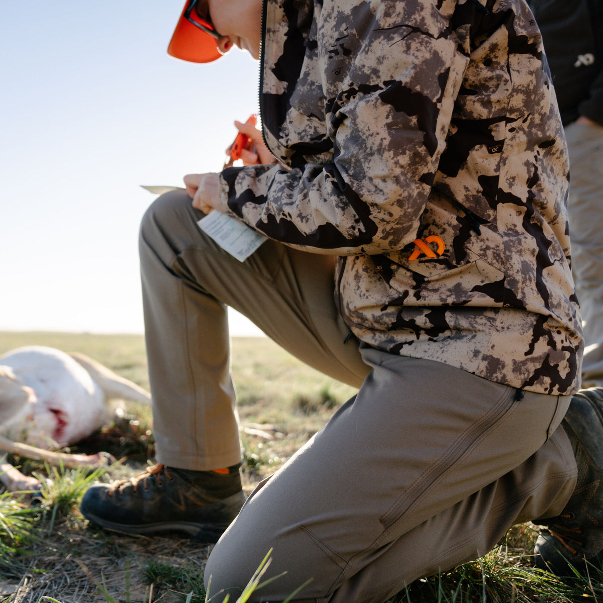 Hunter kneeling outdoors wearing a beige and dark brown camouflage jacket with an orange logo, olive green rugged pants with a vertical zippered thigh pocket, and dark brown hunting boots.
