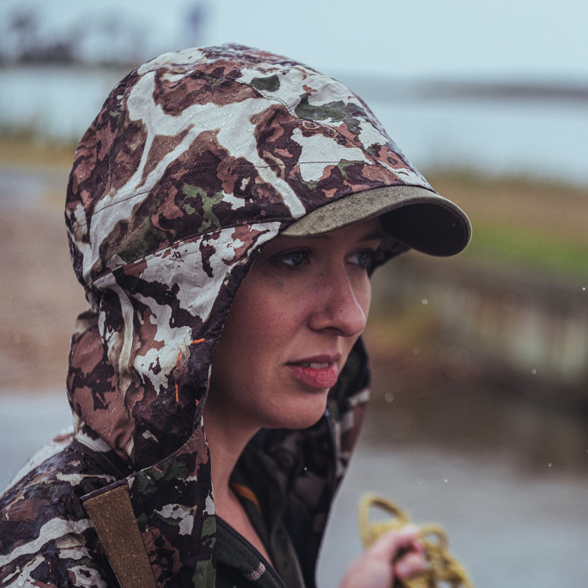 A First Lite brown, green, and white camouflage hooded waterproof jacket with a brim, covered in water droplets.