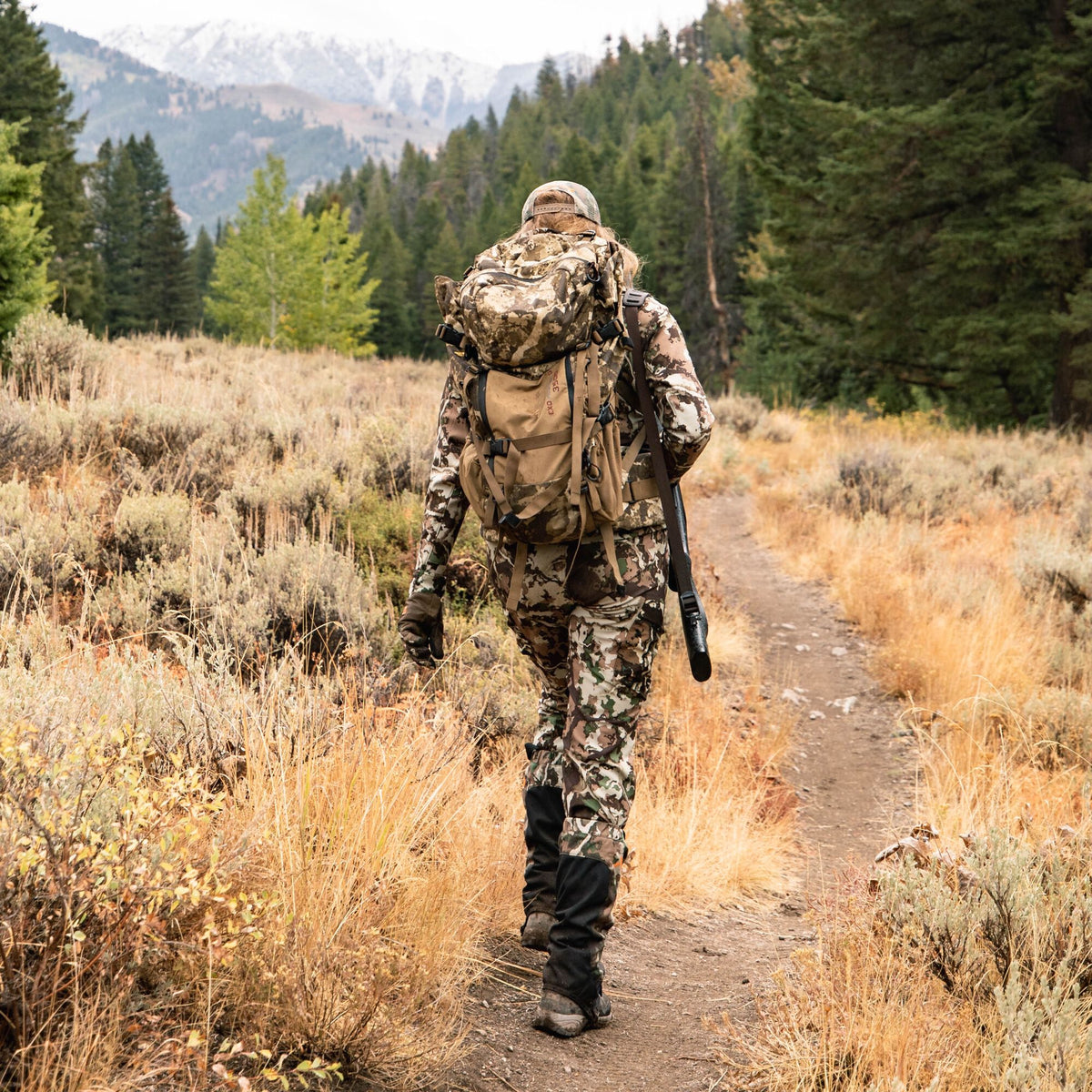 A person in green and brown camouflage hunting gear, with a large backpack and a rifle, walks away on a dirt path.