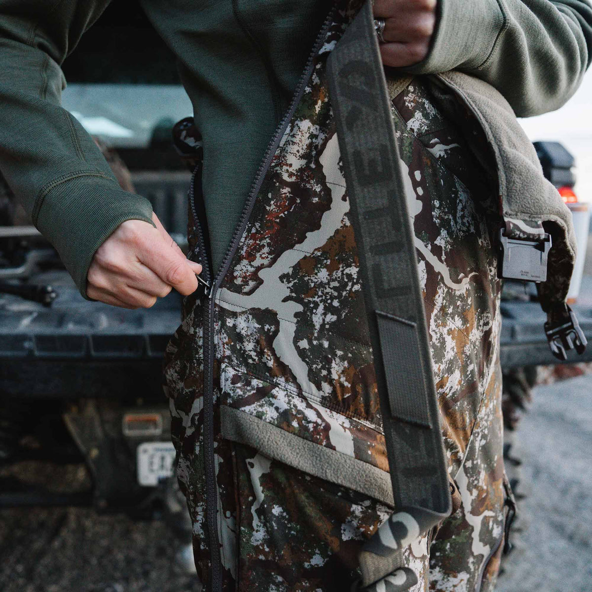 A person zips up mottled brown, green, and white camouflage hunting bib overalls, featuring a visible grey First Lite branded strap.