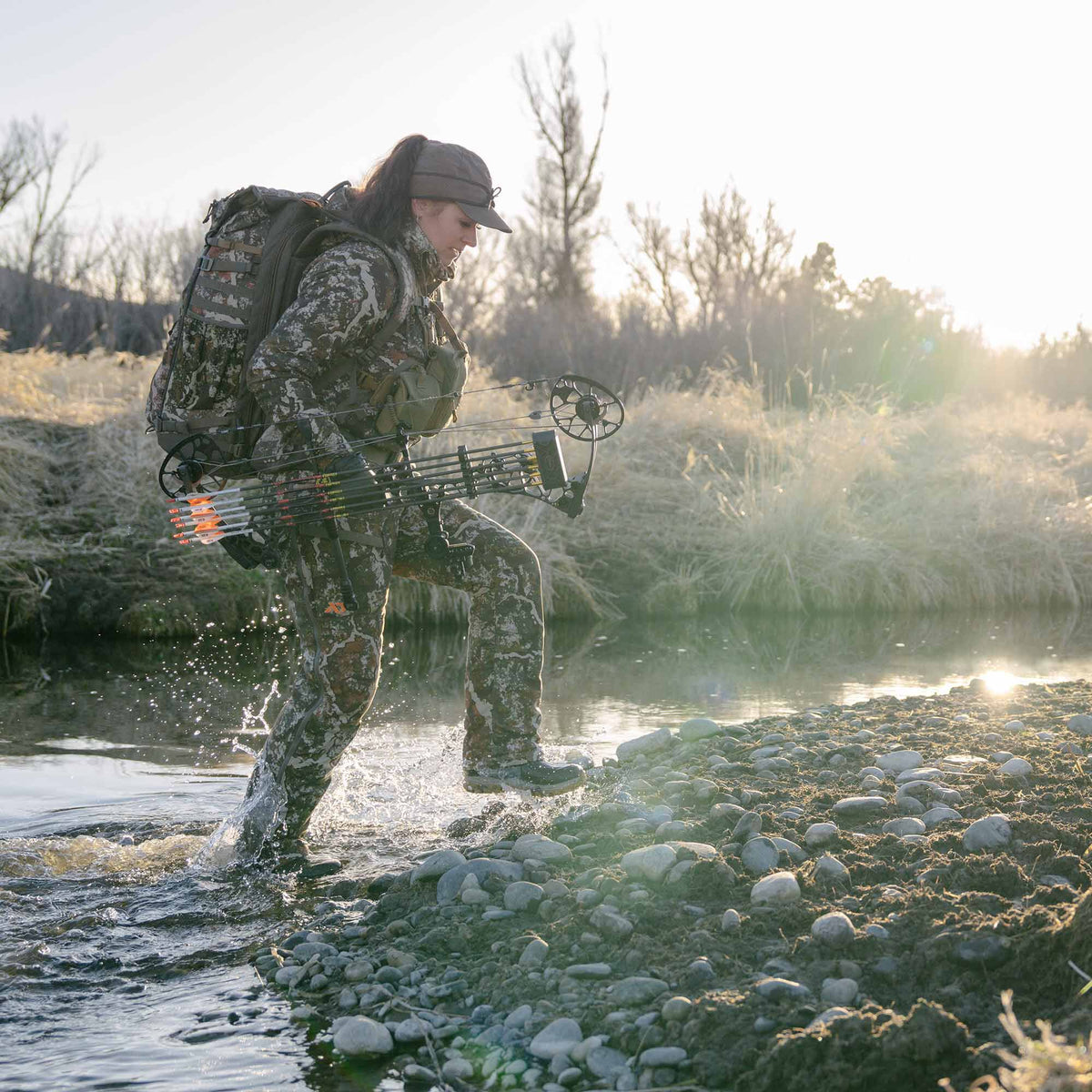 A woman in full camouflage hunting gear, including a backpack and a bow with arrows, wades through a shallow, rocky stream.