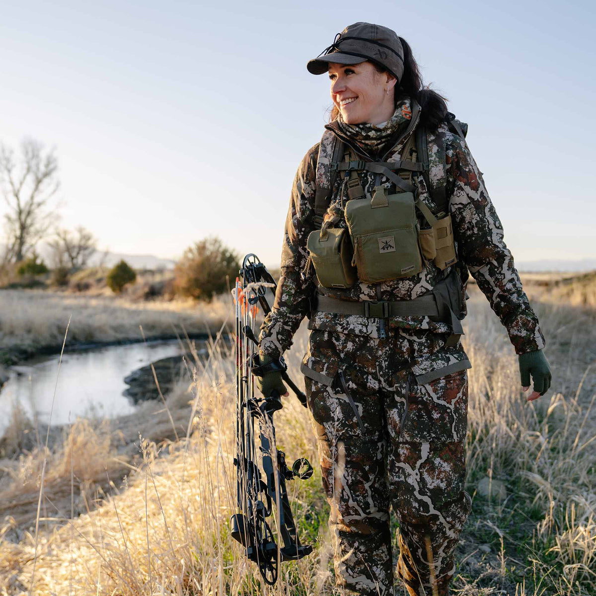 A smiling hunter in a mottled brown, white, and green camouflage suit with an olive chest rig and dark cap holds a black compound bow in a grassy field.