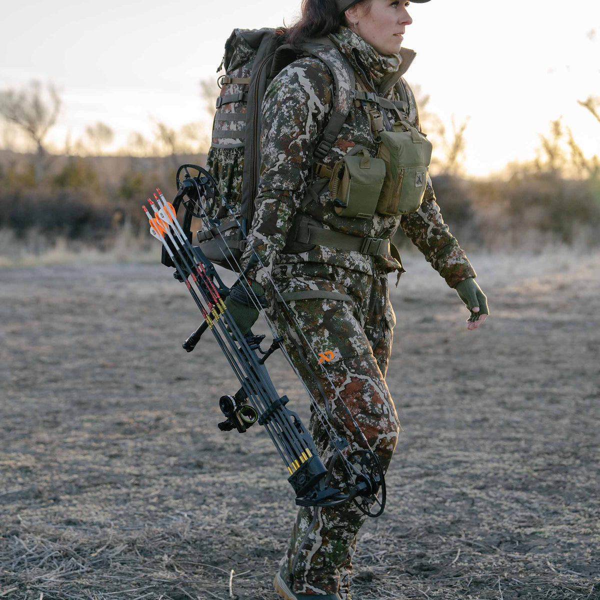 A woman in brown, green, and white camouflage hunting gear, wearing a matching backpack and olive chest rig, carries a black compound bow with white and orange arrows while walking outdoors.