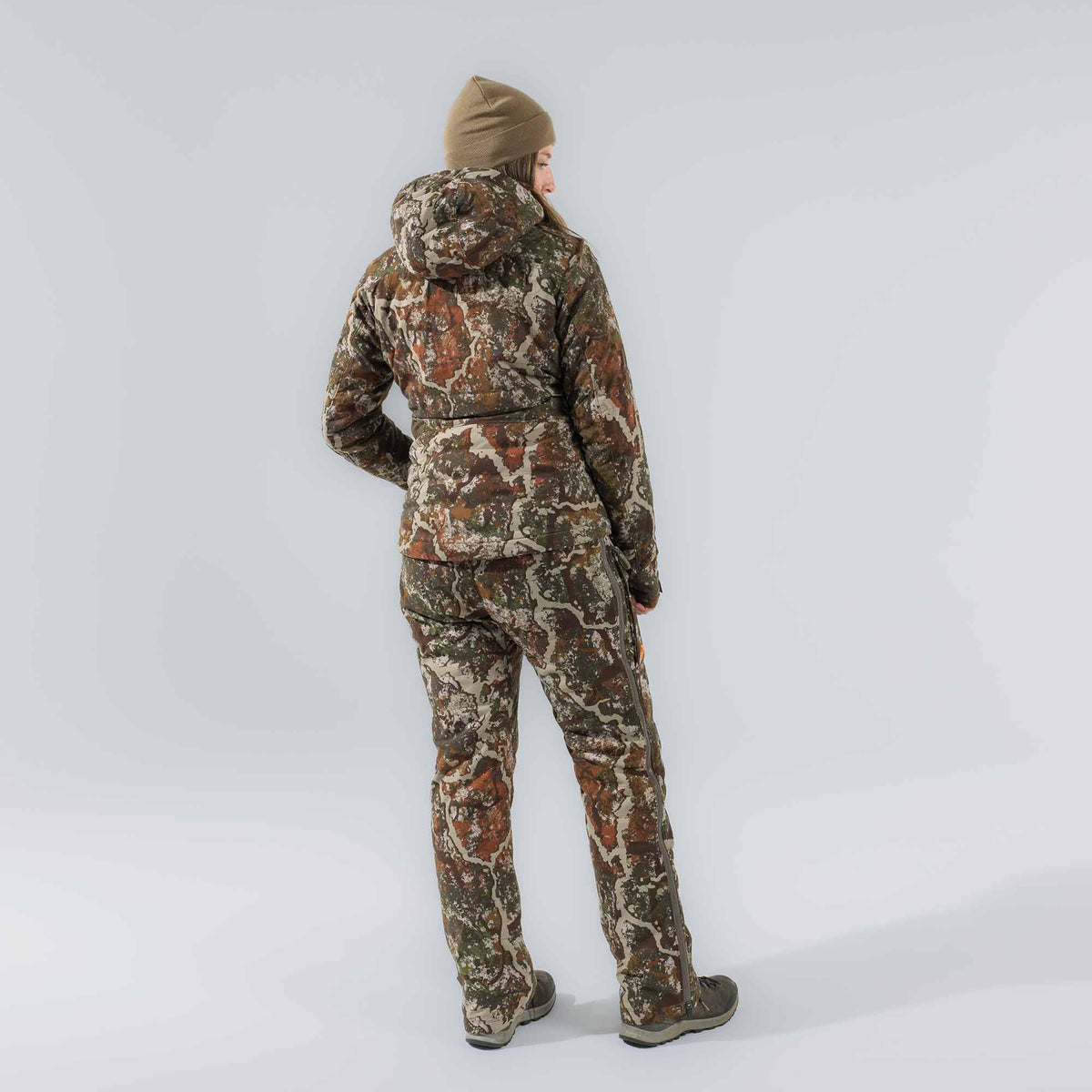 A woman wears a brown, green, and beige camouflage hunting suit with a hooded jacket, waist belt, and full-length zippered pants, seen from behind.