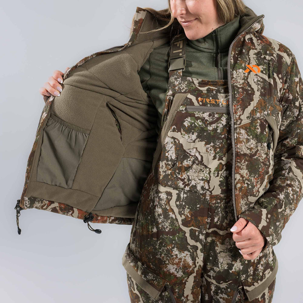 Woman wearing a camouflage jacket open to show its olive green fleece lining and internal pocket, with matching bib overalls underneath.