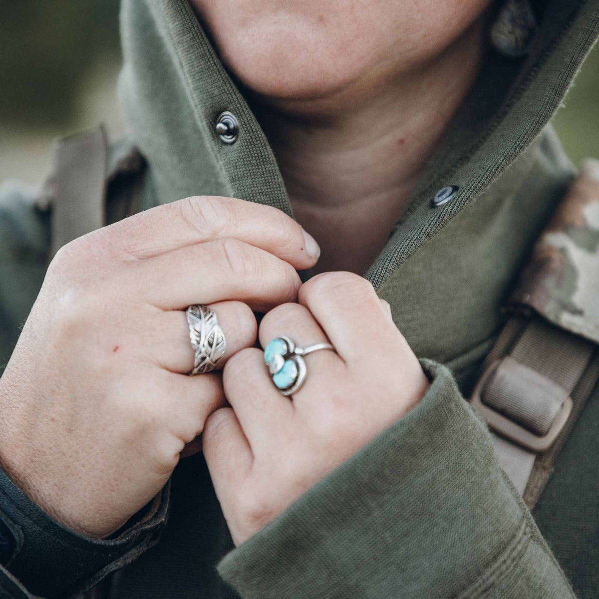 Hands adjust an olive green snap-button top, wearing silver leaf and turquoise rings, with a tan backpack strap visible.