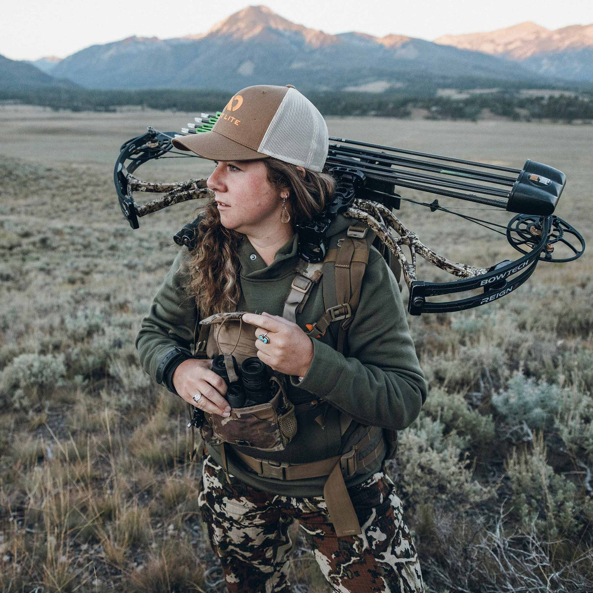 Woman in olive green hoodie, camouflage pants, and tan First Lite cap carries a Bowtech Reign compound bow with arrows and binoculars in a chest harness.