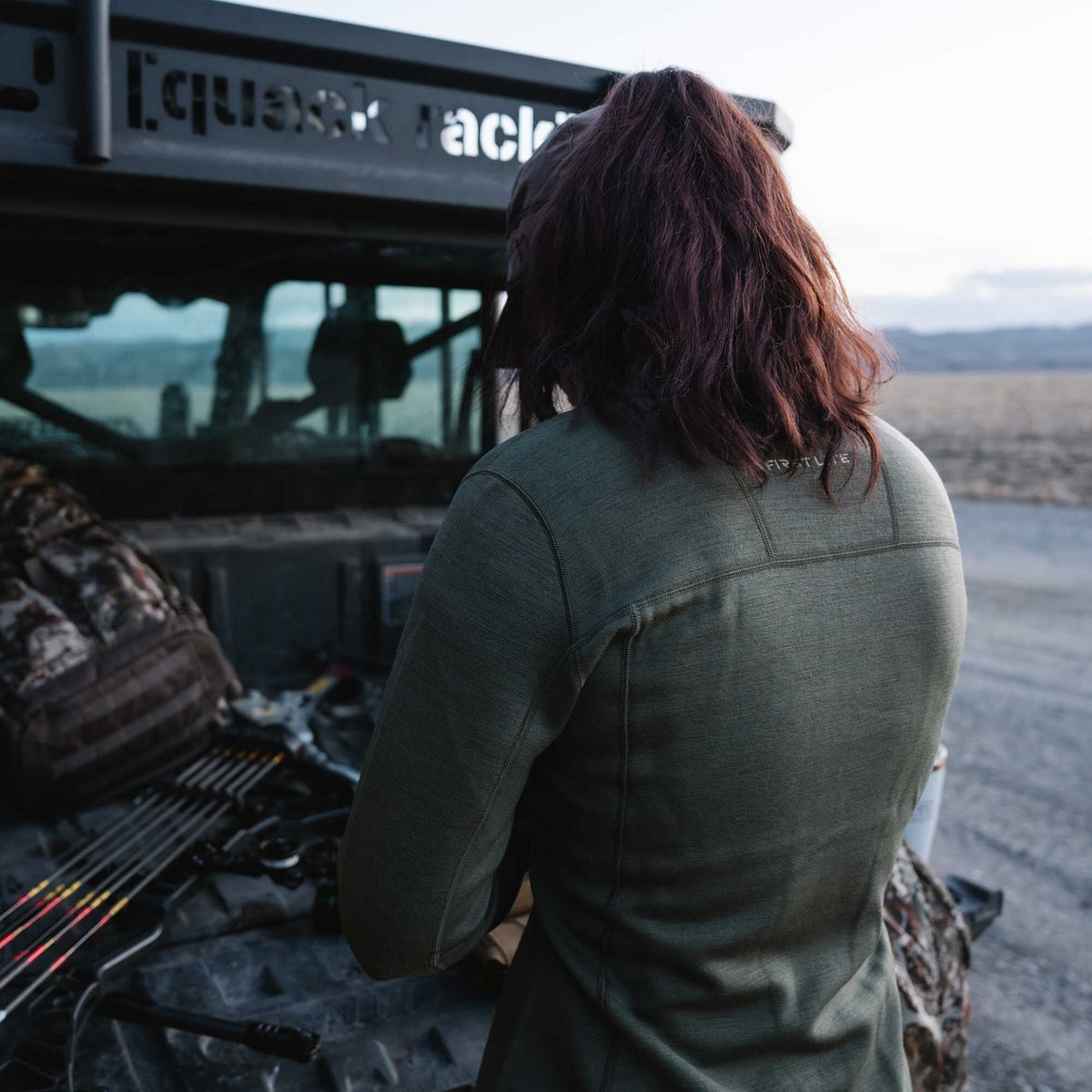 A person wears a dark green First Lite long-sleeved base layer shirt with visible stitching on the shoulders and back.