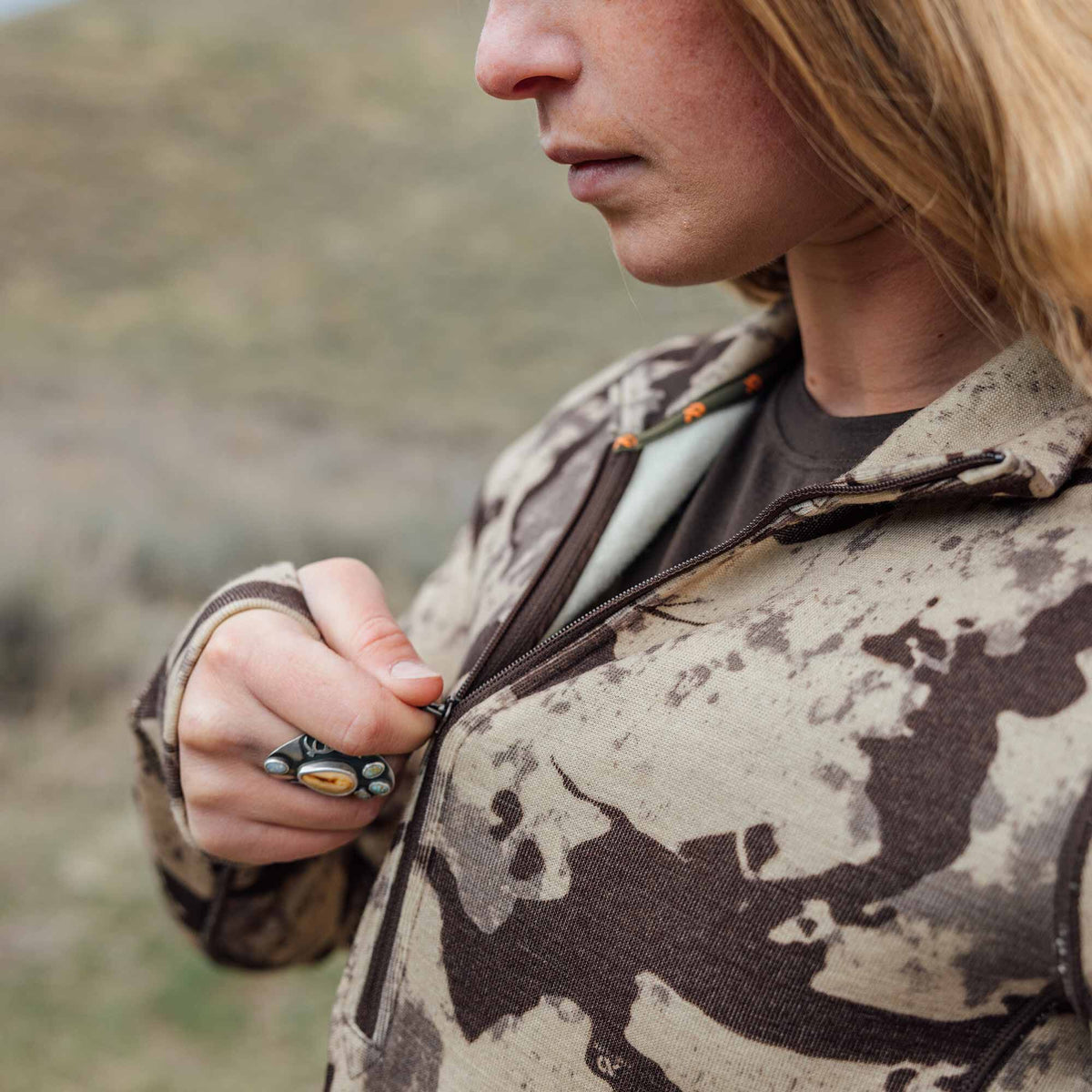 Close-up of a person zipping up a light beige and dark brown camouflage jacket, featuring a decorative ring and an orange zipper pull inside.
