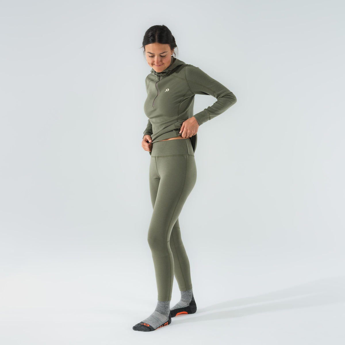 A woman wears an olive green base layer set, featuring a hooded quarter-zip top with a white chest logo and matching high-waisted leggings.