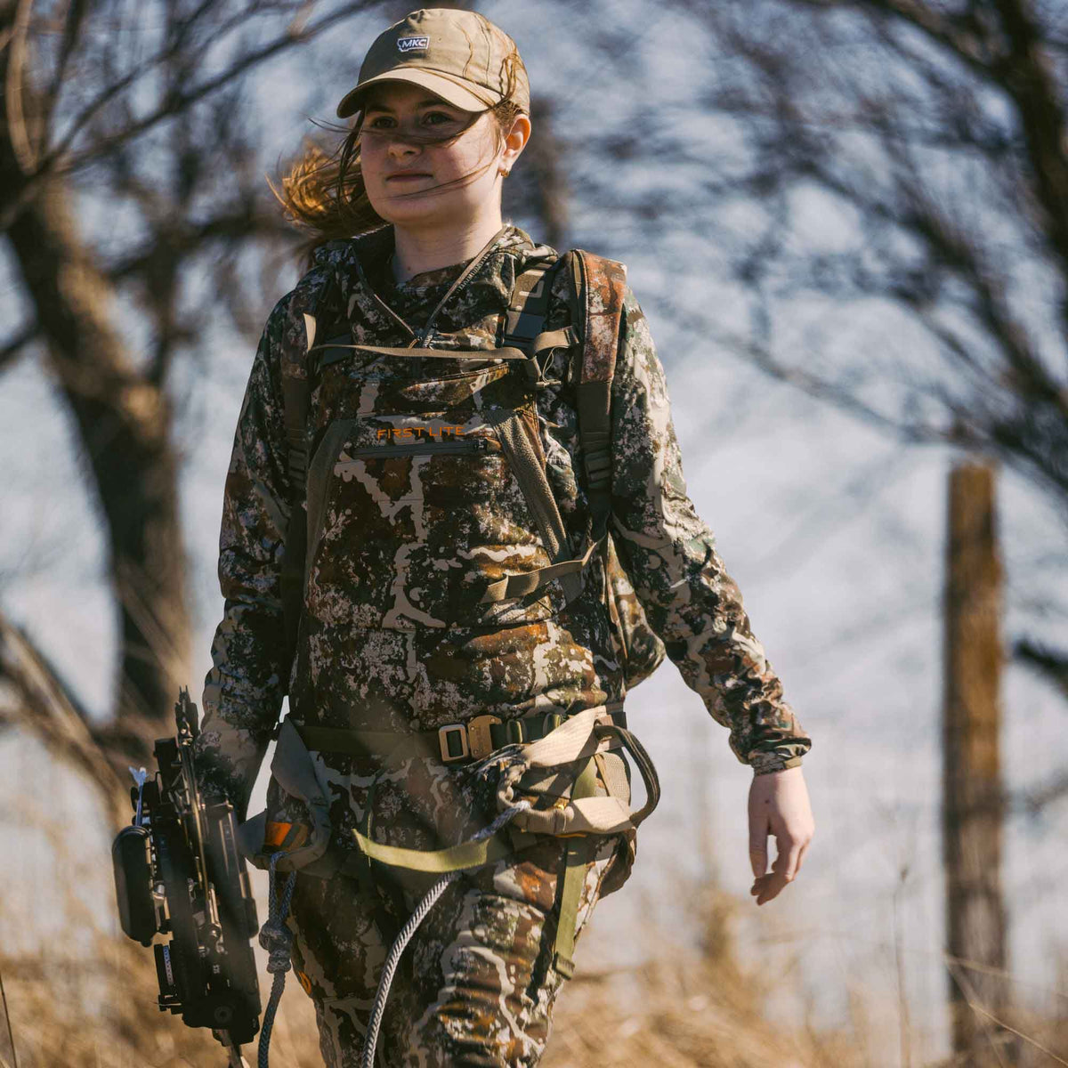 A woman wears a tan MKC baseball cap and a multi-tone camouflage hunting hoodie and pants with a First Lite chest rig, safety harness, and holds a compound bow.