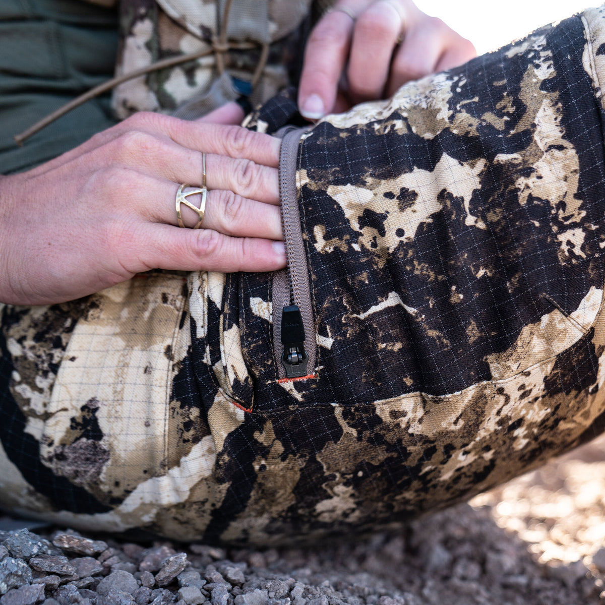 Close-up of a hand zipping up camouflage ripstop pants with dark and light brown patterns and orange accent stitching.