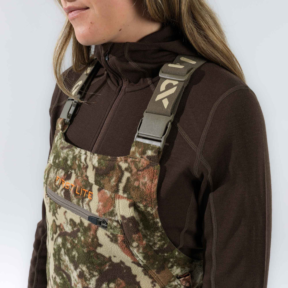 Camouflage hunting bibs featuring an orange 'FIRST LITE' logo, a zippered chest pocket, and adjustable shoulder straps.