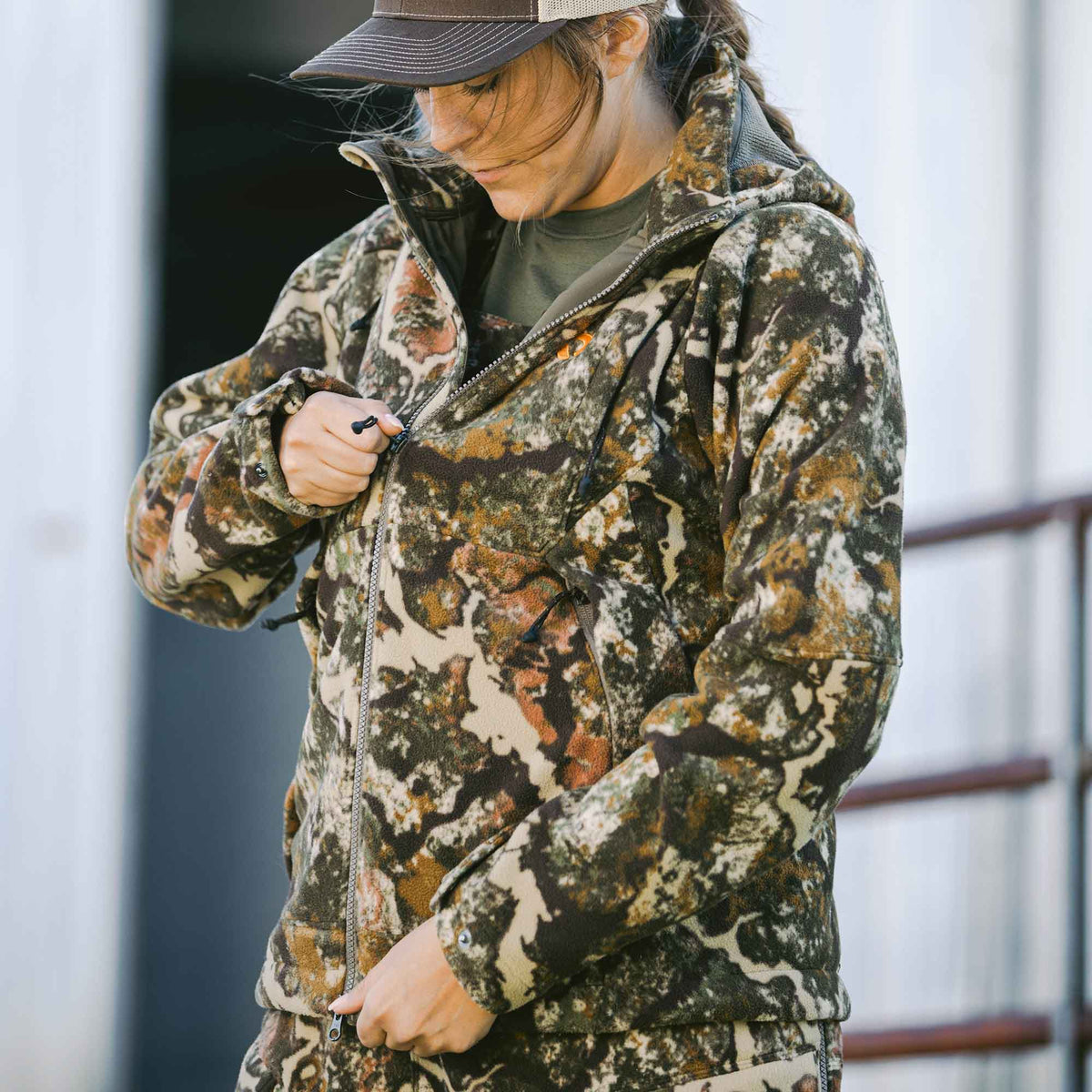 A woman zips up a green, brown, tan, and orange camouflage full-zip hooded fleece jacket.