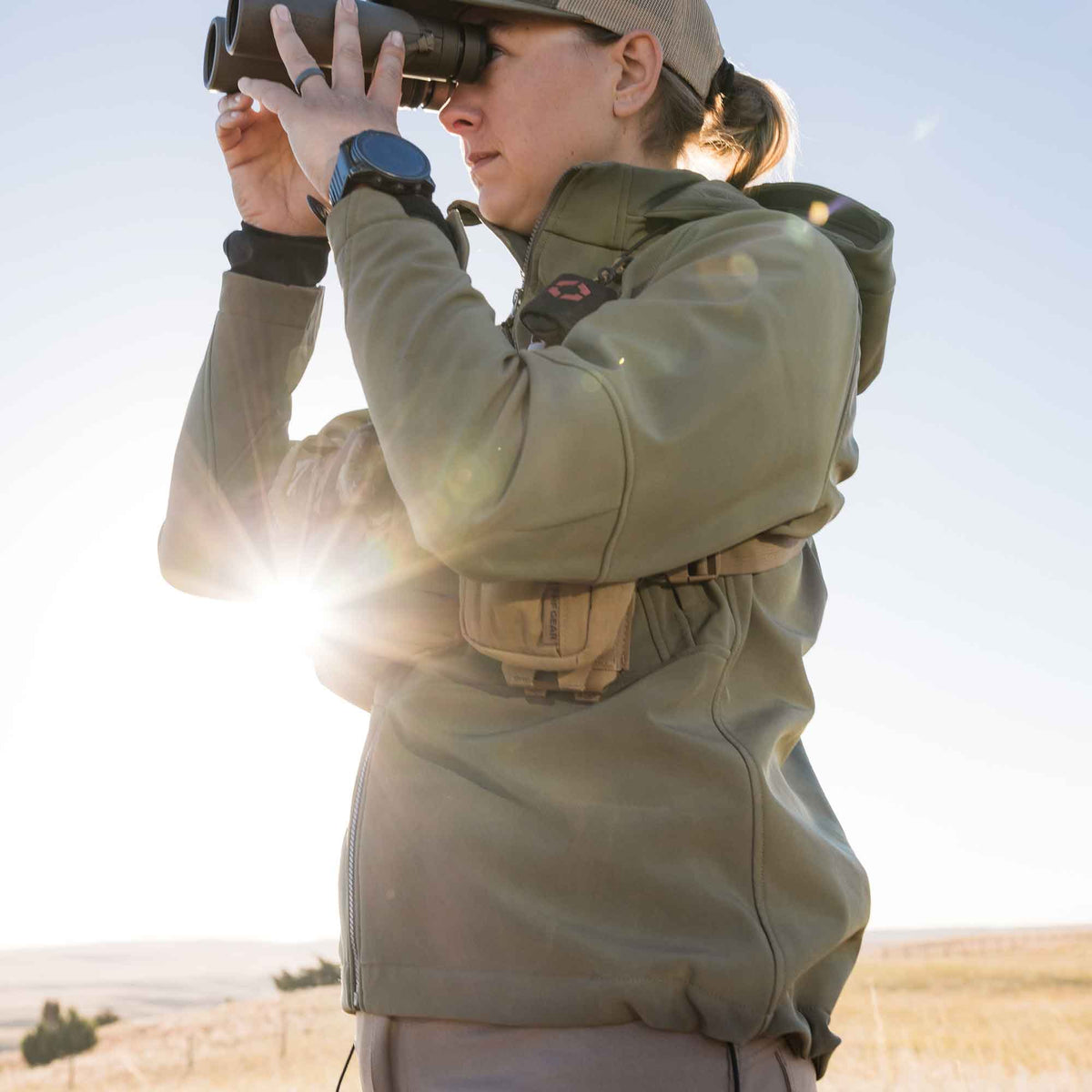 Olive green zippered outdoor jacket on a woman using binoculars, with a tan utility pouch at her waist.