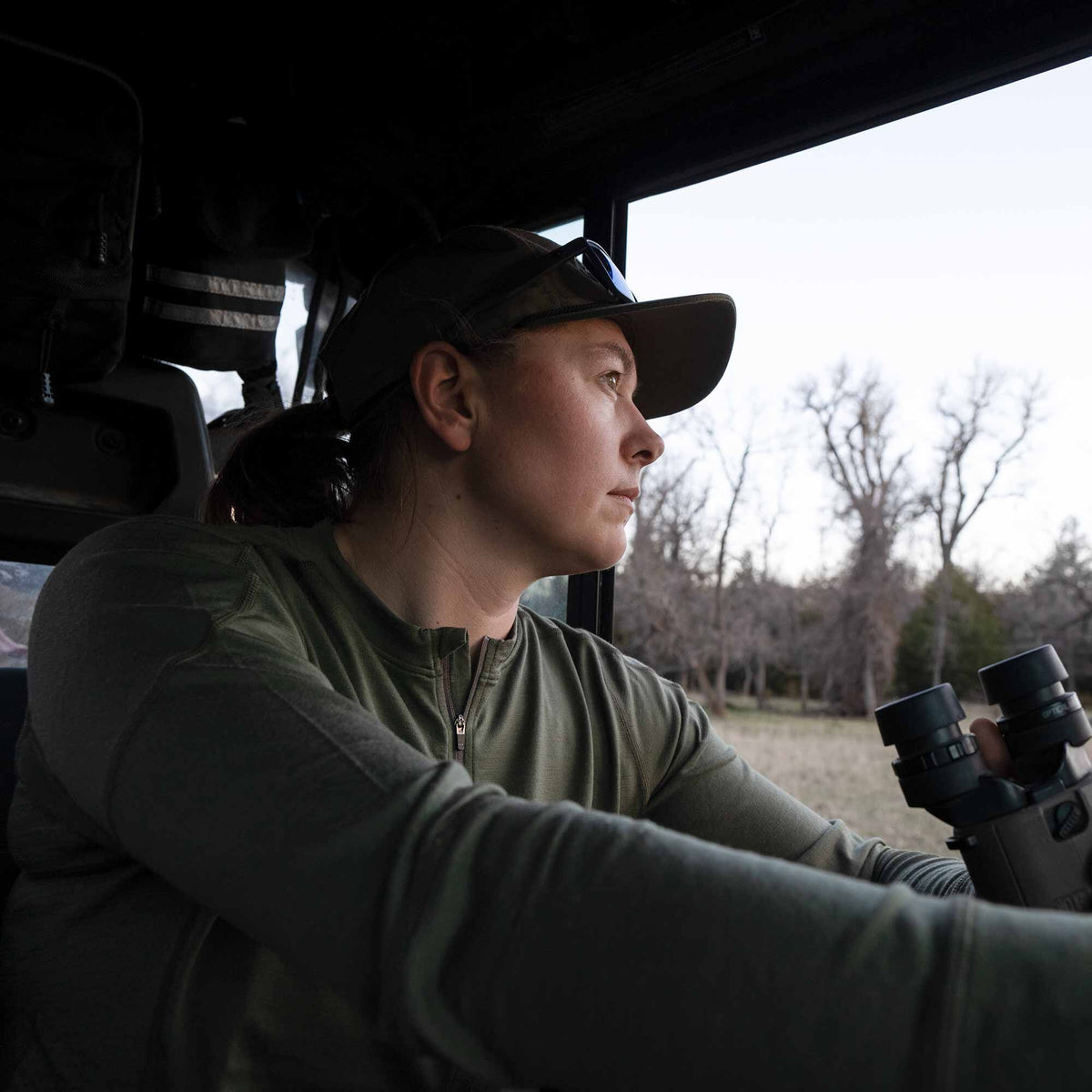 A woman wears an olive green quarter-zip long-sleeve shirt with visible shoulder stitching, looking out from a vehicle while holding binoculars.