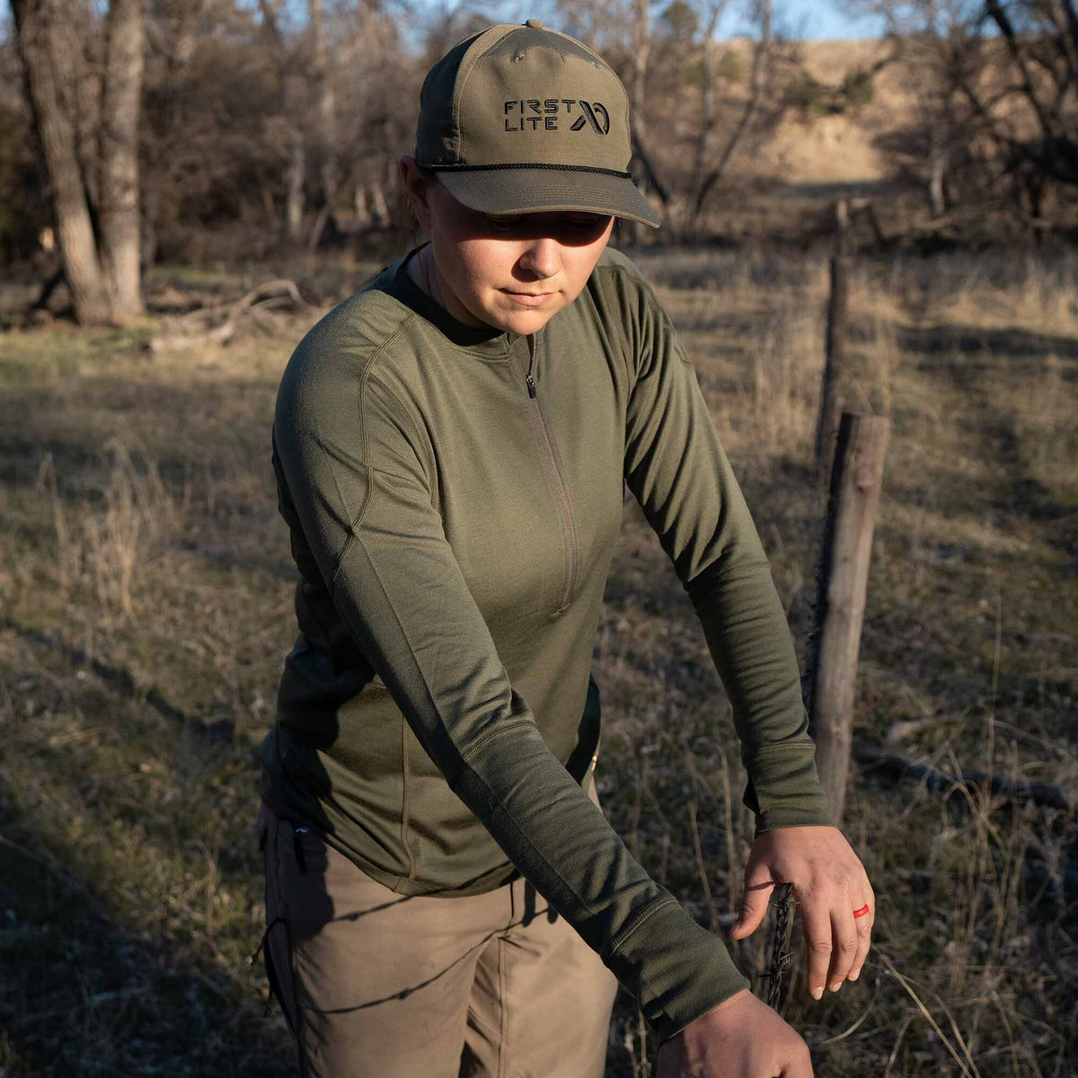 An individual wears an olive green quarter-zip long-sleeve shirt with prominent seams and a matching 'FIRST LITE' baseball cap with a rope detail.