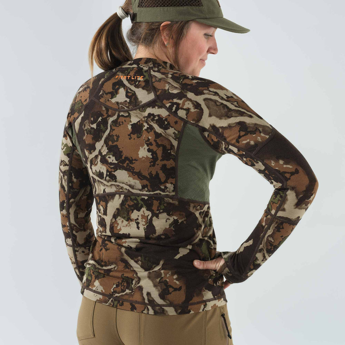 A woman models a brown, tan, and green camouflage long-sleeve base layer shirt featuring green mesh panels and an orange "FIRST LITE" logo on the upper back.