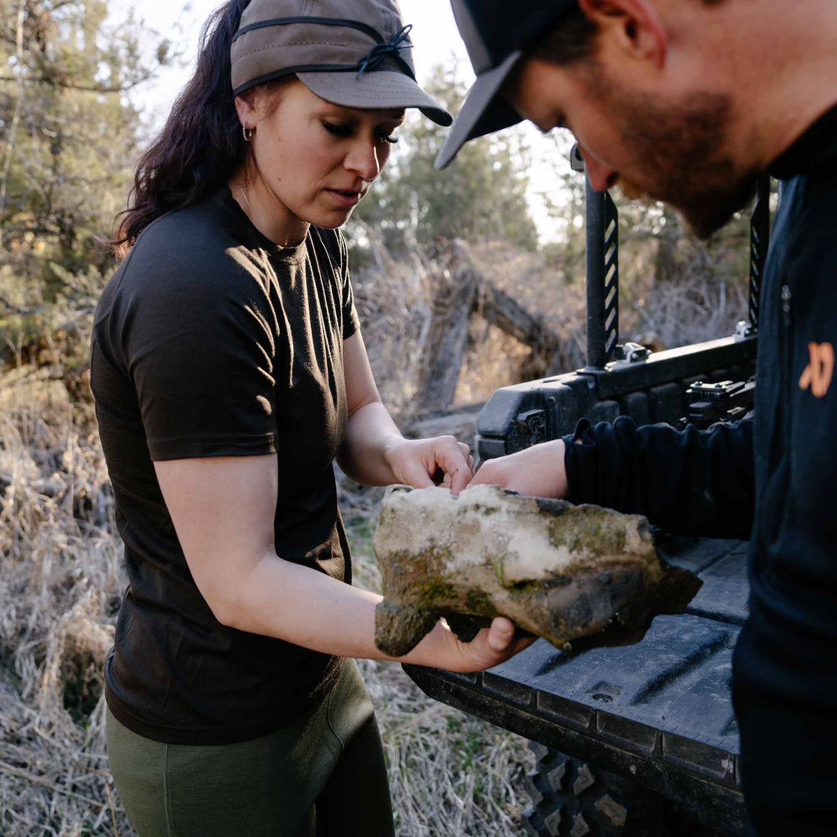 Woman wearing a dark olive green short-sleeve crew neck shirt, examining a mossy rock outdoors.