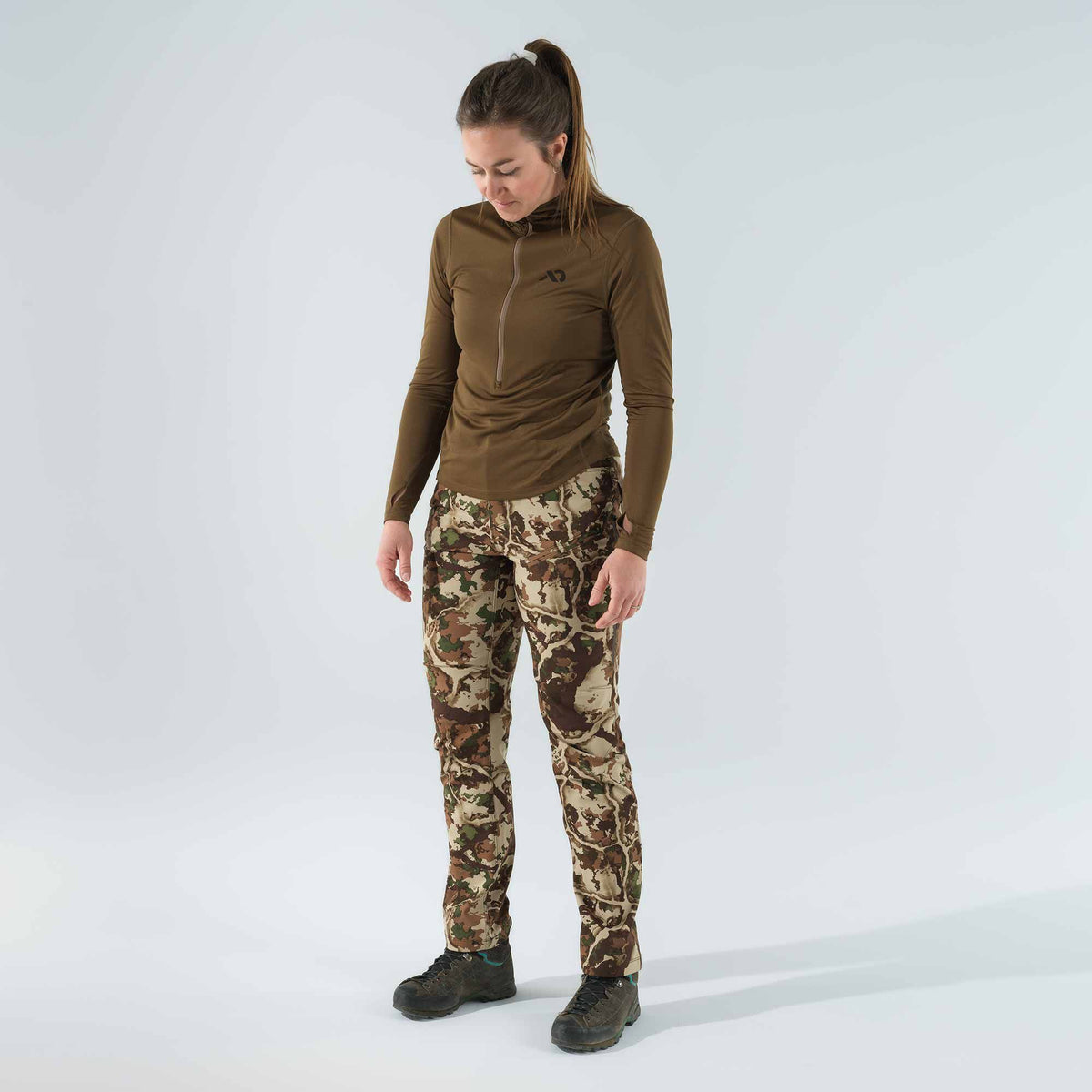 Woman wearing an olive green long-sleeved quarter-zip shirt with thumbholes and camouflage hunting pants.