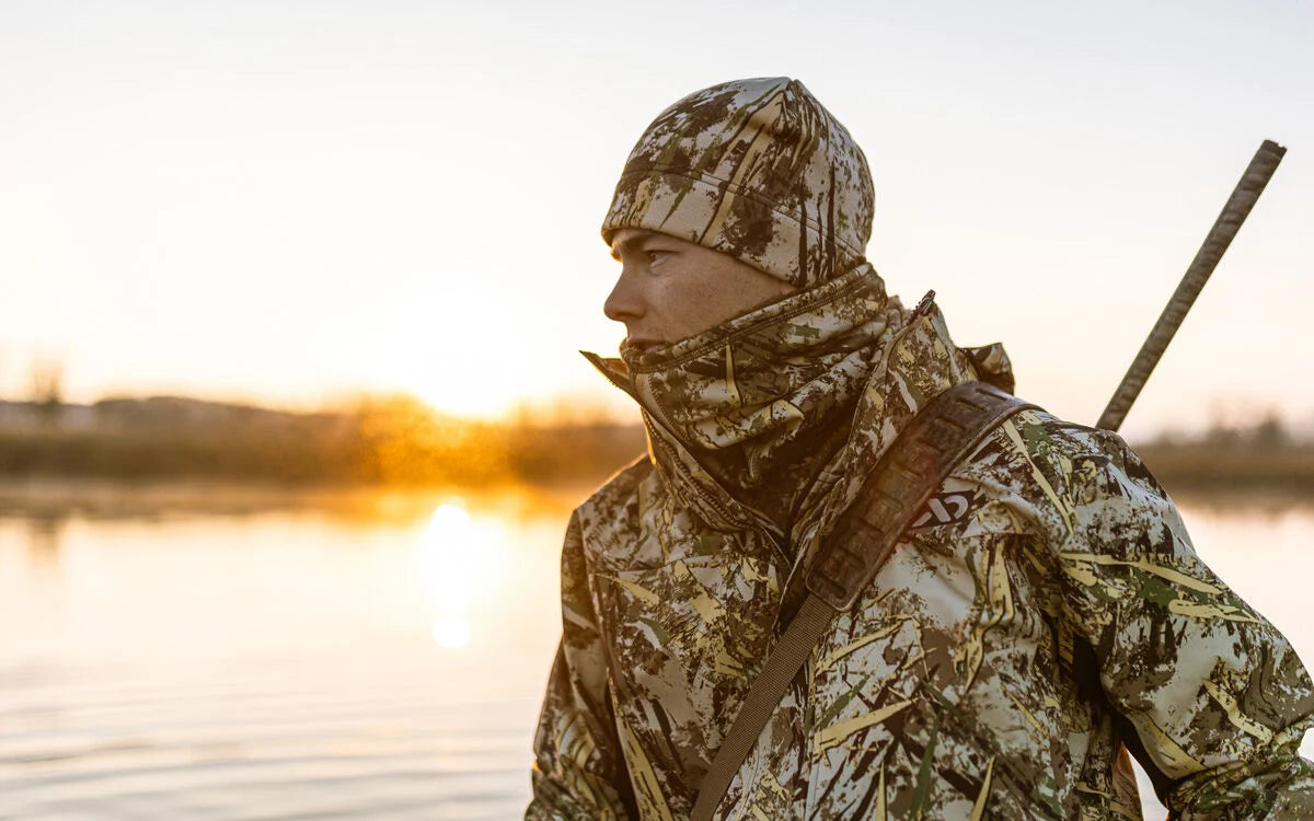 Camo hunting jacket, beanie, and neck gaiter on a hunter with a rifle at dawn.