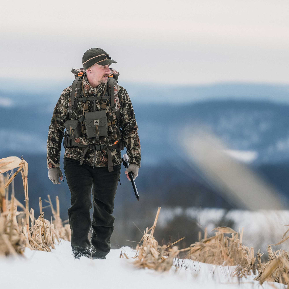 Man walking in the filed wearing camo jacket, dark green cap, light green gloves and dark moss pants.