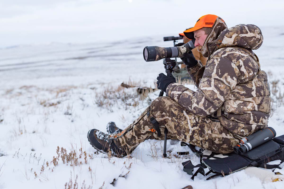 Person in brown camo winter gear, orange hat, using black spotting scope in snow.