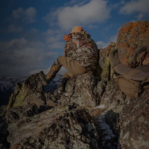 Camo jacket, tan pants, orange hat on a person sitting on a rocky mountain peak.