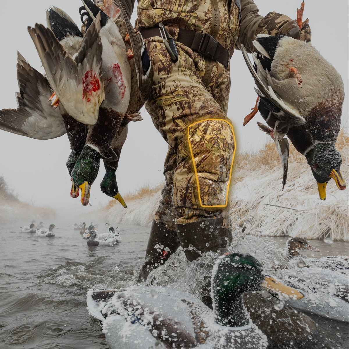 Camo hunting waders with padded knees, worn by a hunter wading through a snowy river.