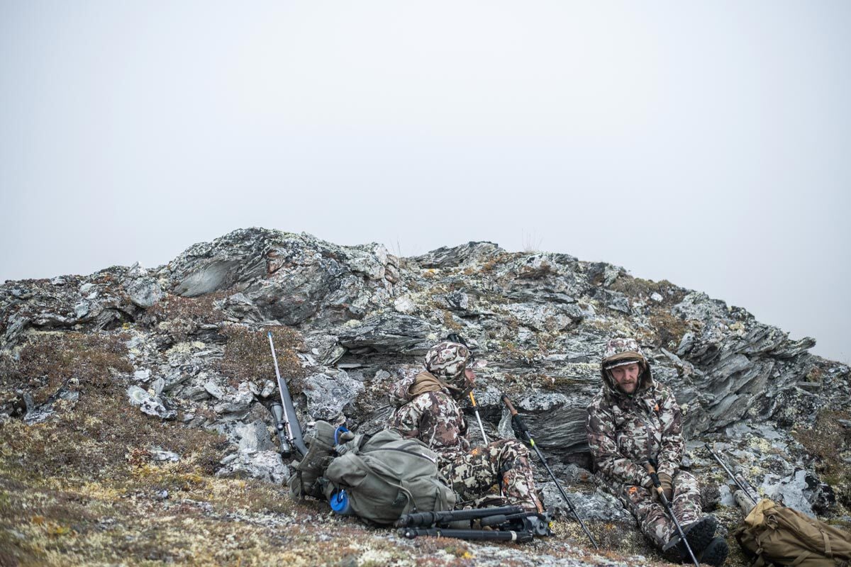 Two hunters in brown camouflage hunting apparel, rifles, and backpacks on rocky terrain.