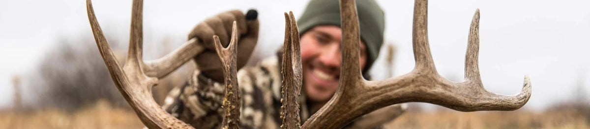 Smiling hunter holds large brown deer antlers; wearing green beanie and camo.