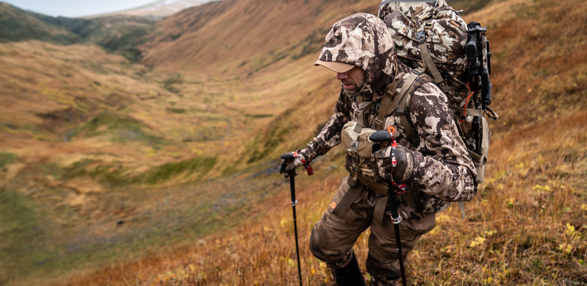 Man in brown camo gear hiking with rifle-attached pack and trekking poles.