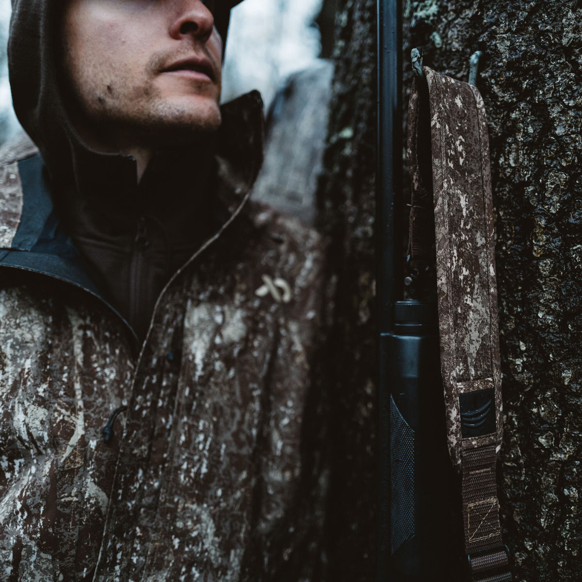 Brown camouflage rifle sling with a black logo accent attached to a dark rifle, beside a person in a matching camo jacket.
