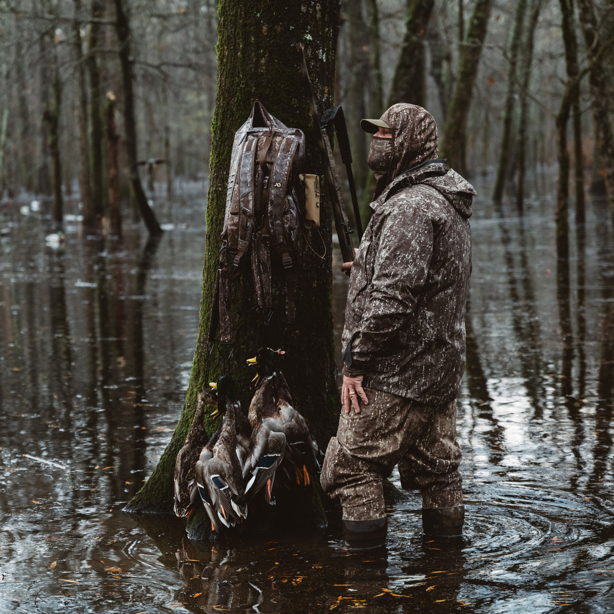 A man wears mottled brown camouflage hunting waders, a hooded jacket, and a face mask while standing in flooded woods next to a tree with a matching backpack.