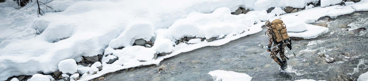 Hunter in camo with tan backpack and trekking pole wading through icy river.
