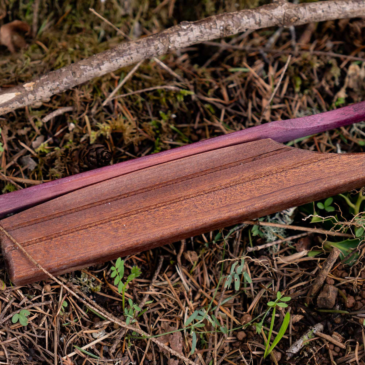 Dark reddish-brown wooden bow riser with carved grooves and a purple wooden bow limb on a forest floor.