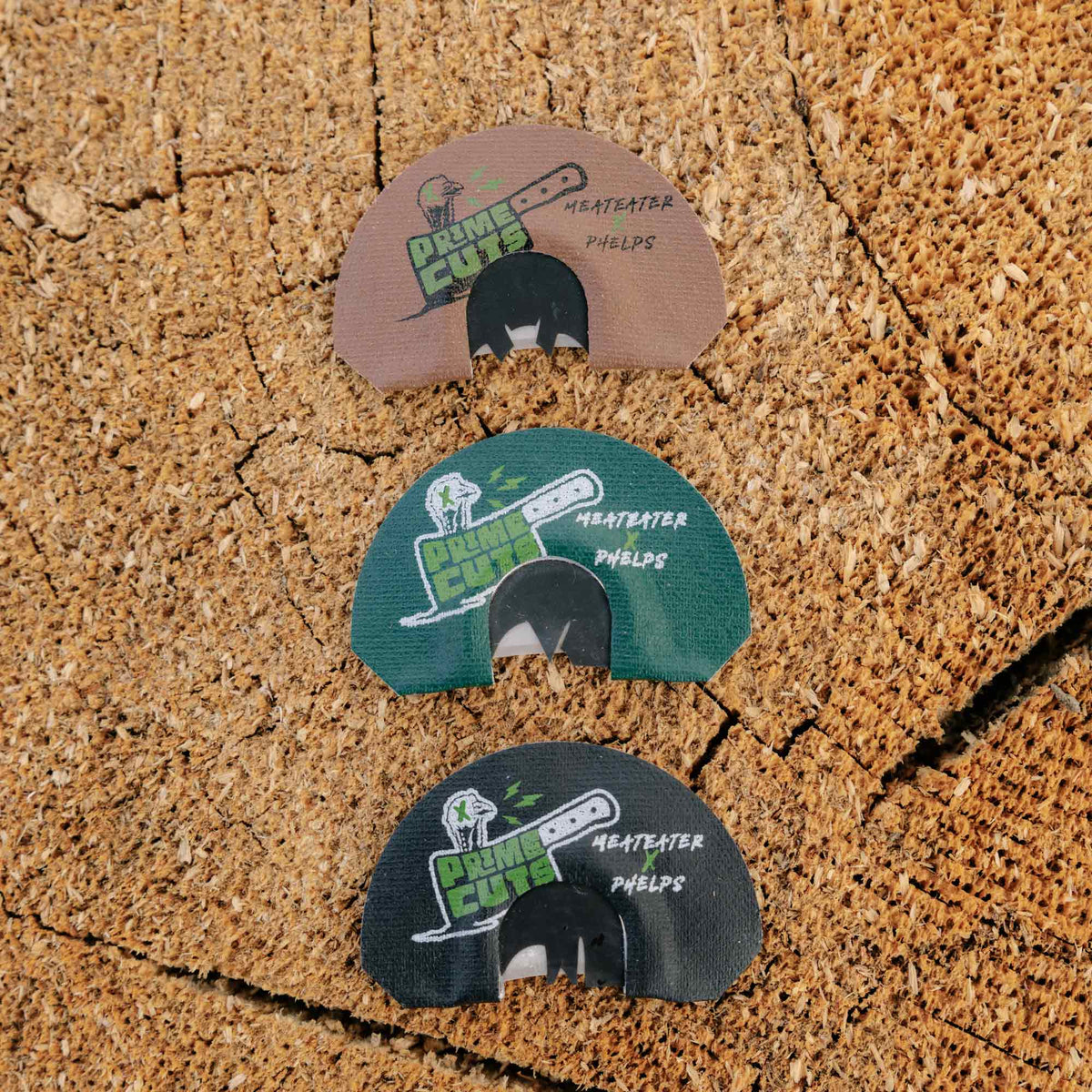 Three Phelps Meateater Prime Cuts turkey diaphragm calls in brown, green, and black, each with a cleaver logo, arranged vertically on a wood surface.