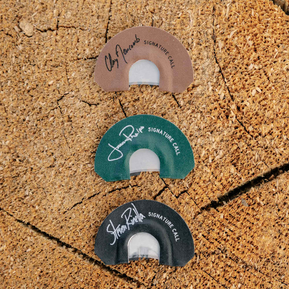 Three semi-circular hunting diaphragm calls in brown, green, and black, each with a white signature and "SIGNATURE CALL" text, arranged vertically on a wood surface.