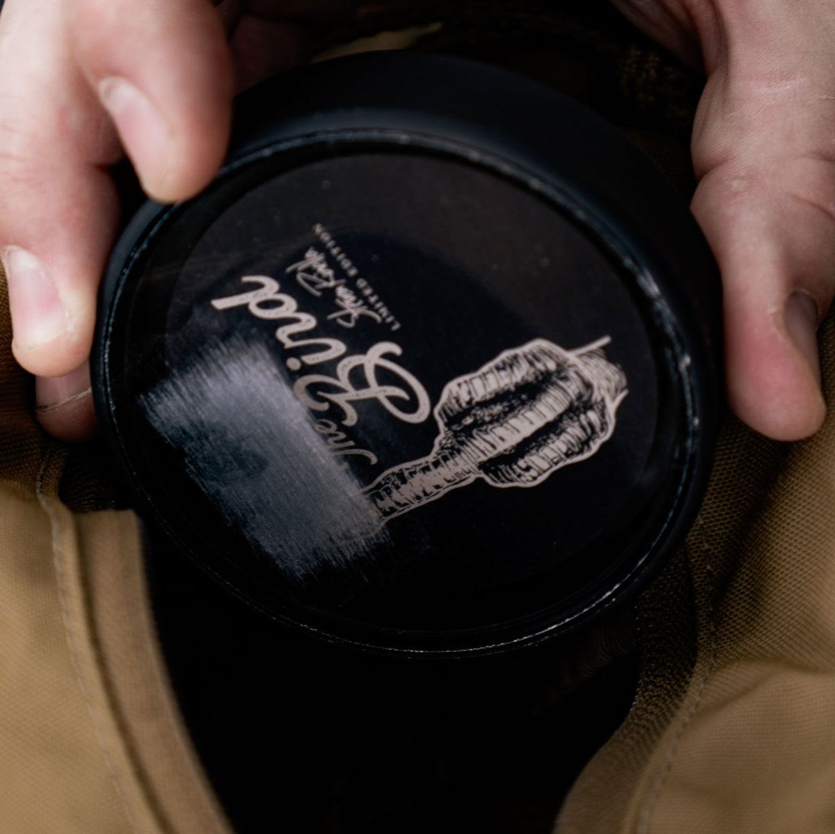 A person's hands hold a round black The Bind Limited Edition tin featuring a logo of a hand holding a quill on its lid.
