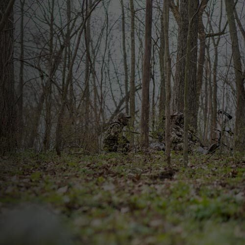 Camouflaged hunters and blind with bow in forest, wearing woodland camo.