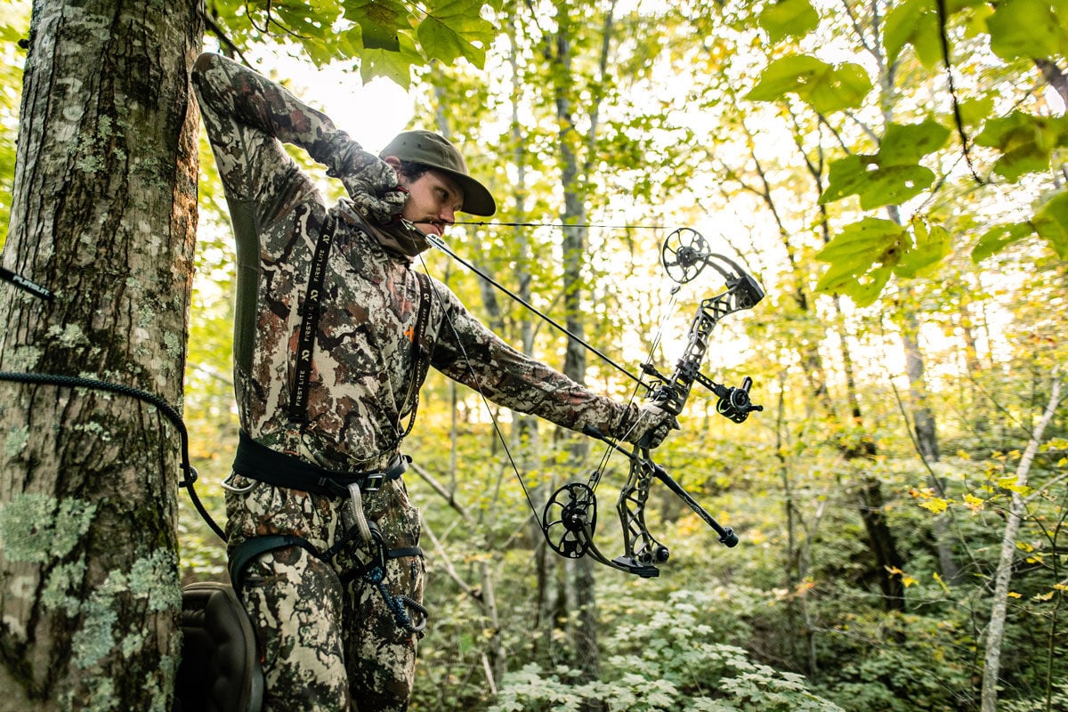 Man in camouflage hunting suit & harness aims compound bow.