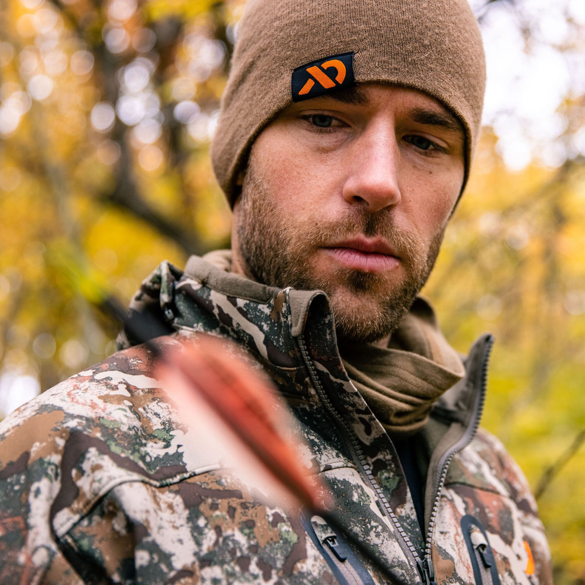 A man wears a brown knit beanie with an orange logo and a zippered camouflage jacket.