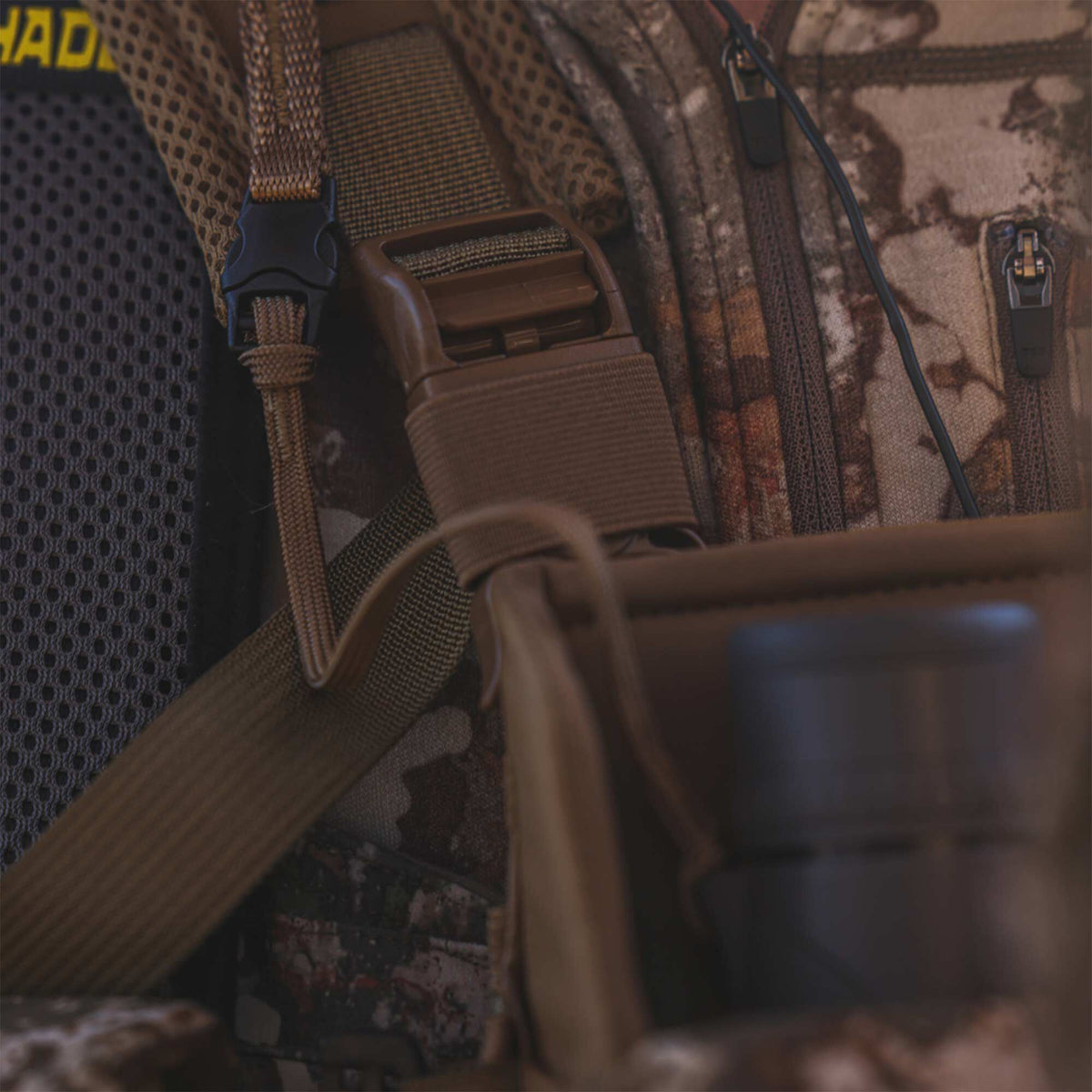 Close-up of a brown and green camouflage backpack featuring tan webbing, black and brown buckles, dark gray mesh padding, and zipper pulls.