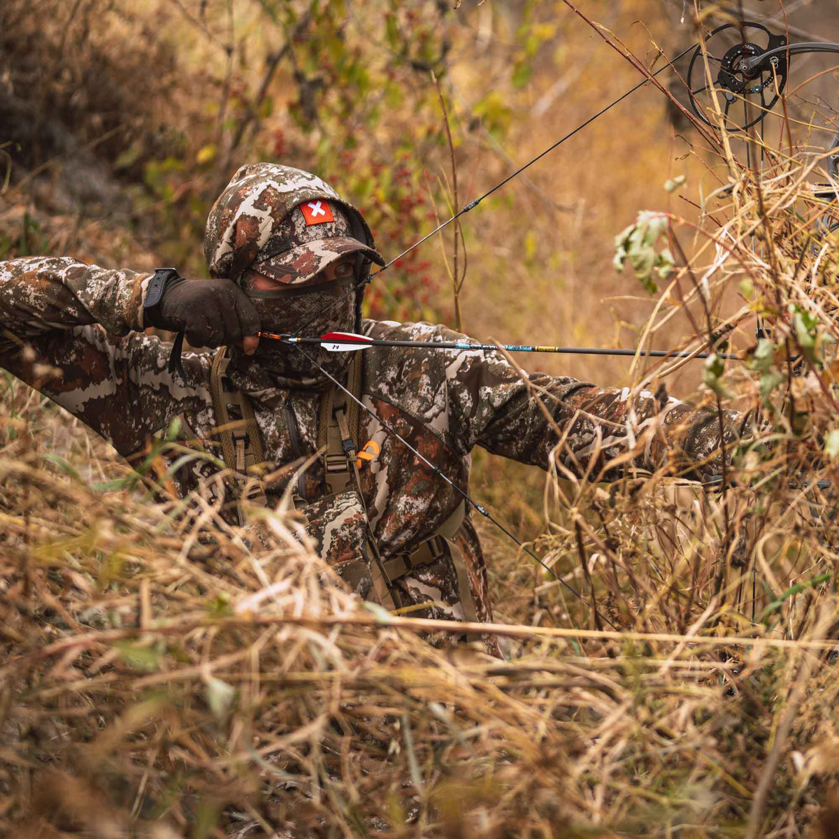 A person in brown and white camouflage gear, including a hat with a red cross, aims a black compound bow with a nocked arrow through tall dry brush.