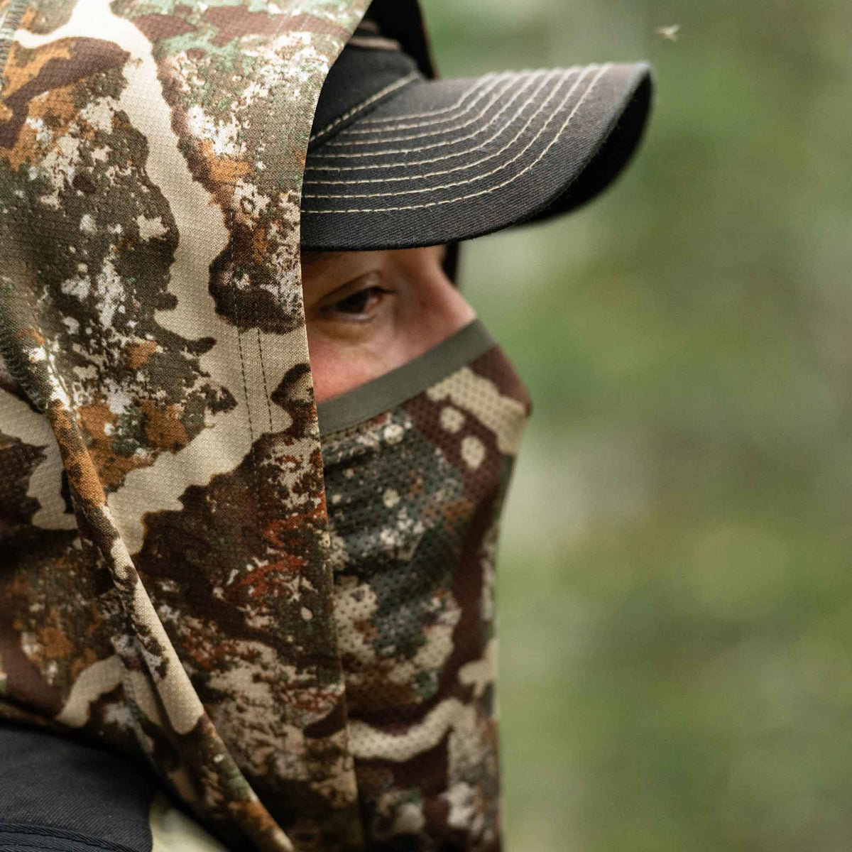 A person wears a multi-color camouflage hooded face mask with mesh, worn over a black cap, showing one eye.