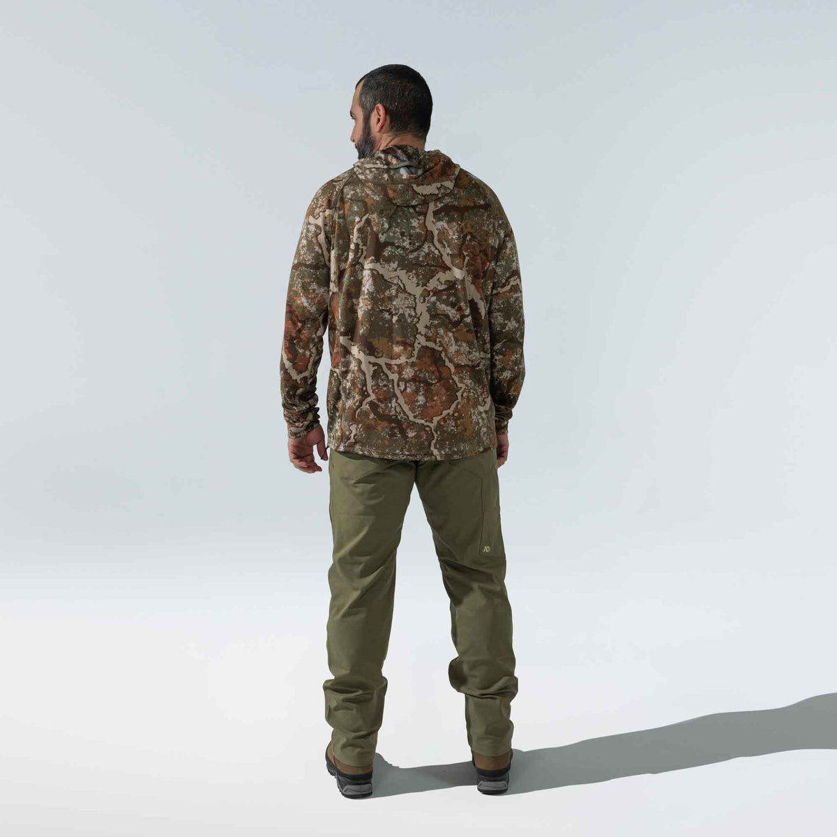 A man seen from behind wears a brown, green, and tan tree-bark camouflage hooded long-sleeve shirt and olive green cargo pants with a small logo on the right thigh.
