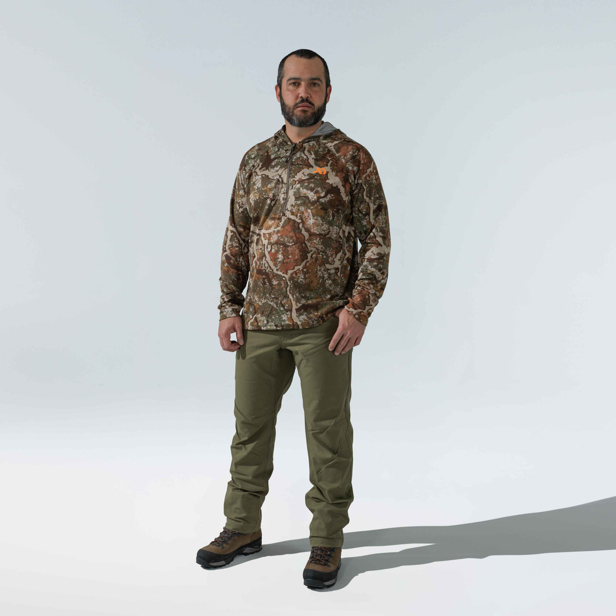 Man wears a camouflage quarter-zip long-sleeve shirt with an orange logo, olive green outdoor pants featuring a zippered thigh pocket, and brown hiking boots.