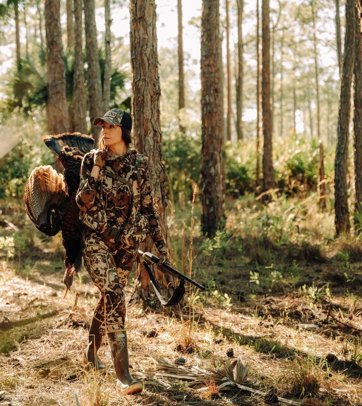 Earth-toned camouflage hunting outfit on woman carrying turkey and shotgun.
